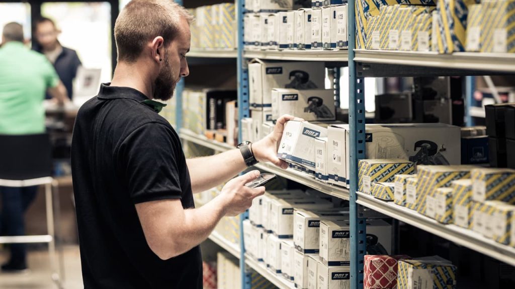 Parts distributor selecting a spare part from a shelf while consulting a code on his smartphone, using innovative automotive solutions for ITGs, parts distributors and wholesalers