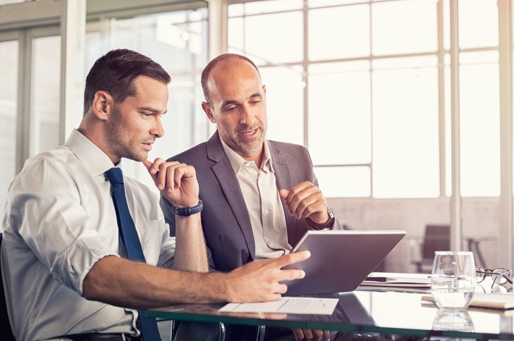 Deux hommes assis côte à côte à un bureau qui réfléchissent ensemble à un projet sur mesure pour réseaux et grands comptes en consultant leur tablette