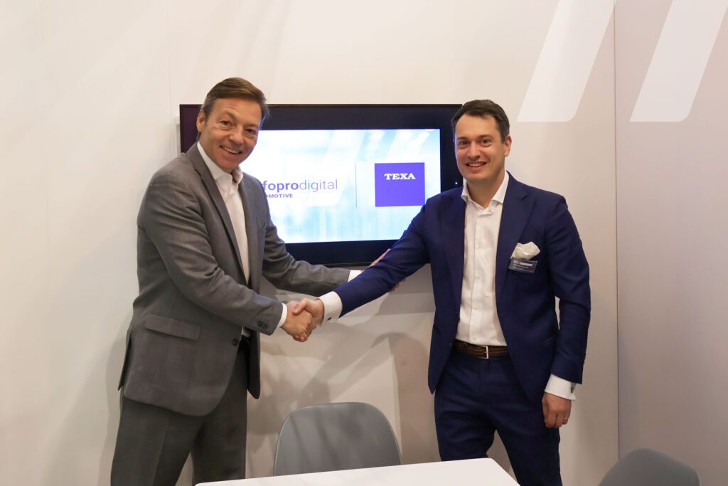 Two representatives of Infopro Digital Automotive and TEXA shaking hands in front of a screen displaying the companies’ logos.