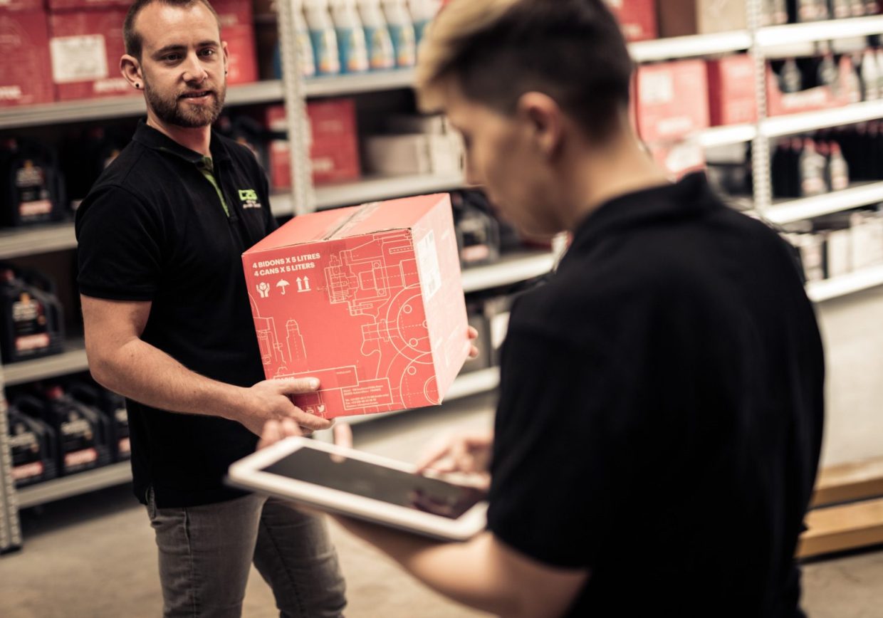 Two parts workers in a distribution warehouse hold boxes of parts while using a tablet catalogue, representing automotive solutions for ITGs, parts distributors and wholesales