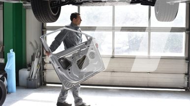 Mécanicien carrossier portant une pièce de carrosserie en atelier dans le cadre d'une intervention comprenant des pièces de réemploi automobiles
