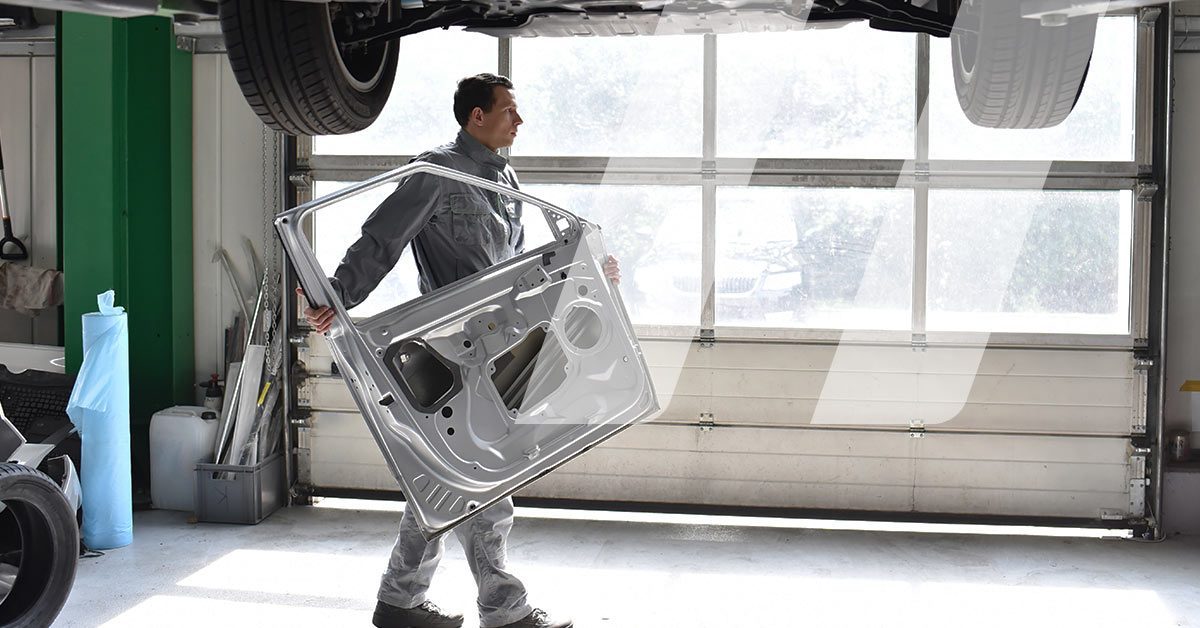 Mécanicien carrossier portant une pièce de carrosserie en atelier dans le cadre d'une intervention comprenant des pièces de réemploi automobiles